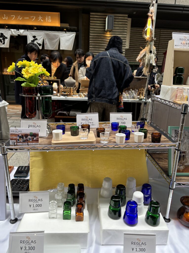 "REGLASのイベント販売ブース全景。ガラス薬瓶やダッペングラスなど色とりどりのガラス雑貨が並び、背景には来場者で賑わうOLD NEW MARKETの様子が写る。